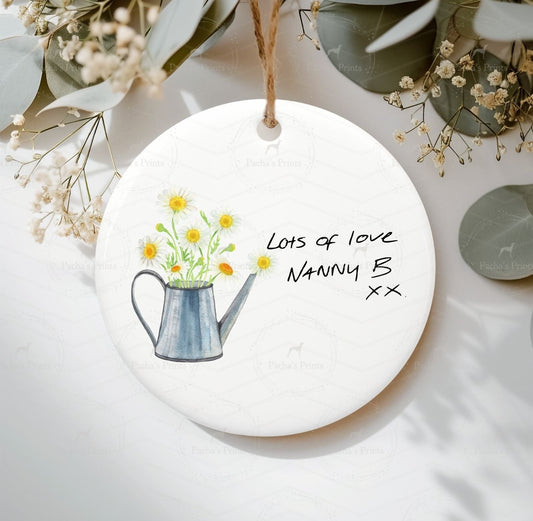 Round ceramic ornament with a watering can and flowers illustration, text on a white background.