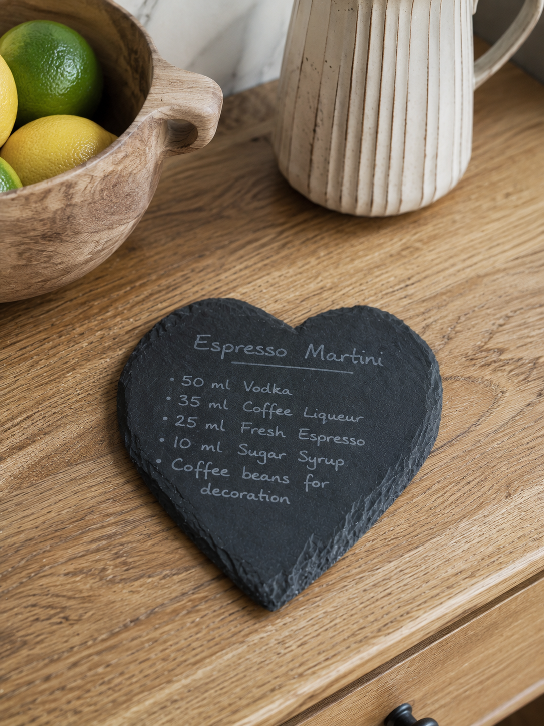 Heart-shaped slate coaster with espresso martini recipe on a wooden surface with lemons and a mug.