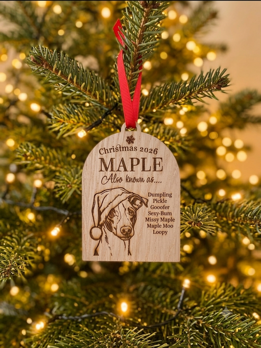 A personalized wooden Christmas ornament hanging from a pine tree branch with warm string lights in the background. The ornament is engraved with a dog portrait and the text 'Christmas 2026 MAPLE Also known as.