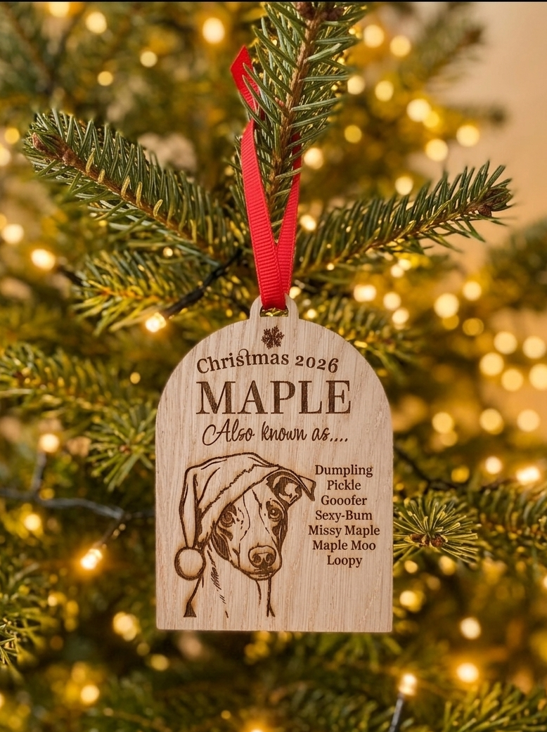 A personalized wooden Christmas ornament hanging from a pine tree branch with warm string lights in the background. The ornament is engraved with a dog portrait and the text 'Christmas 2026 MAPLE Also known as.