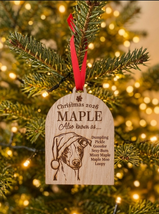 A personalized wooden Christmas ornament hanging from a pine tree branch with warm string lights in the background. The ornament is engraved with a dog portrait and the text 'Christmas 2026 MAPLE Also known as.