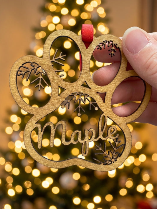 A hand holds a personalized wooden paw-shaped Christmas ornament engraved with the name 'Maple' and small leaf designs, with a blurred Christmas tree and warm lights in the background.
