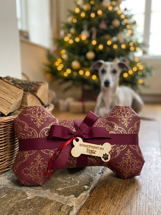 A bone-shaped dog toy wrapped in burgundy damask-patterned paper with a matching ribbon and bow, featuring a wooden tag that reads 'SOMETHING TO Taste'. A Whippet dog sits in the background in front of a decorated Christmas tree.
