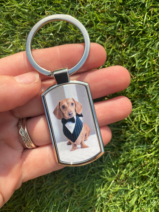 A silver metal keychain with a custom printed photo of a dachshund wearing a tuxedo, held in a person's hand against a background of green grass.
