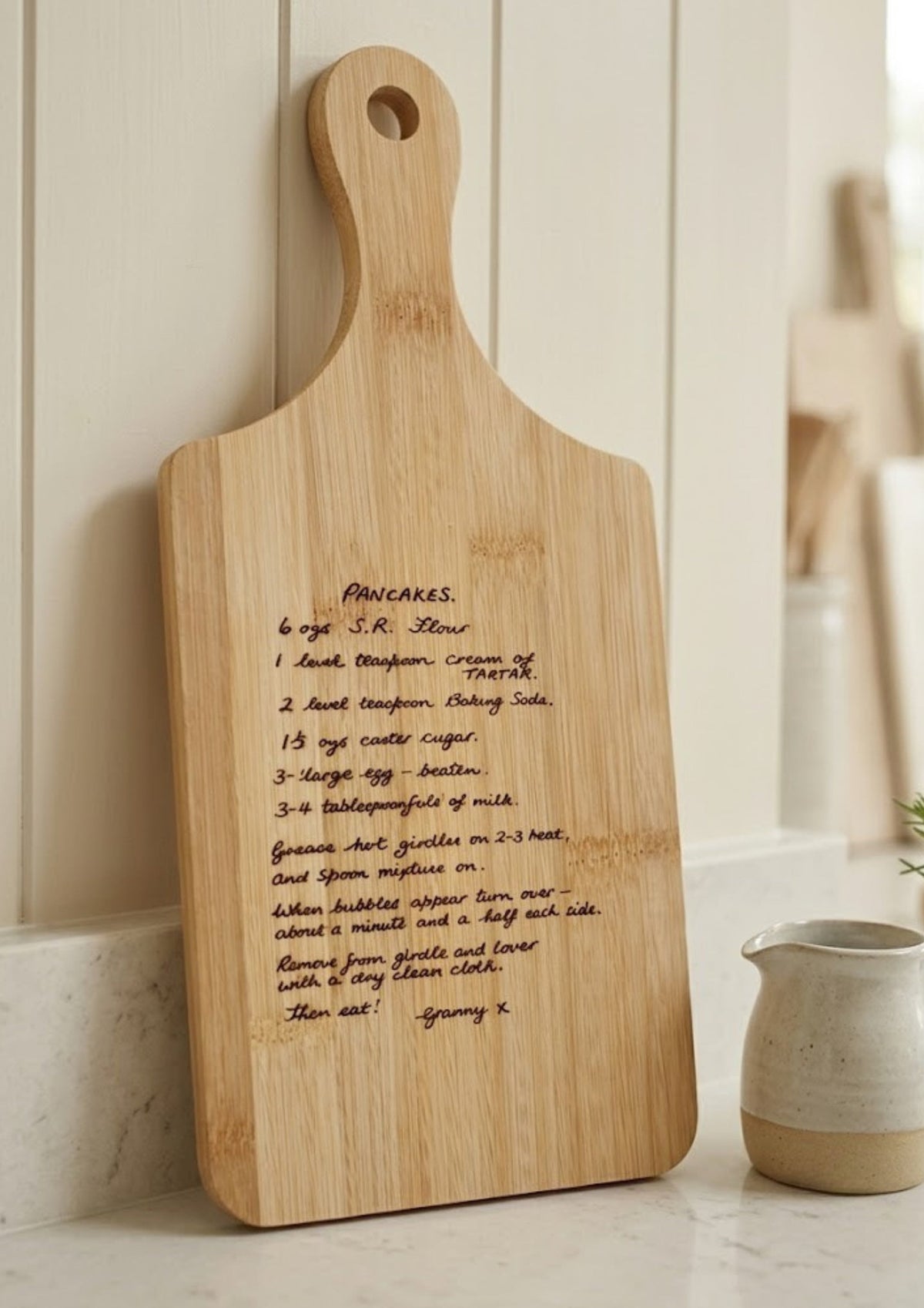 A light natural wood serving board with a handle, featuring a handwritten recipe engraved on the front, leaning against a white paneled wall next to a small cream-colored ceramic pitcher.