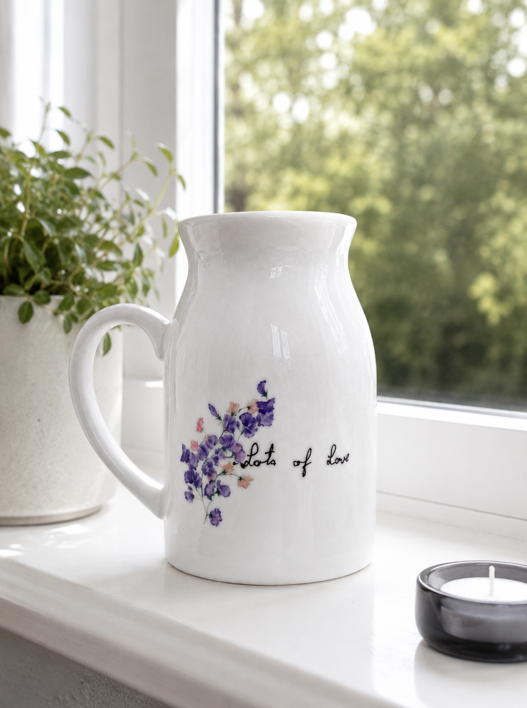 White pitcher with floral design and text on a windowsill with plants and a candle.
