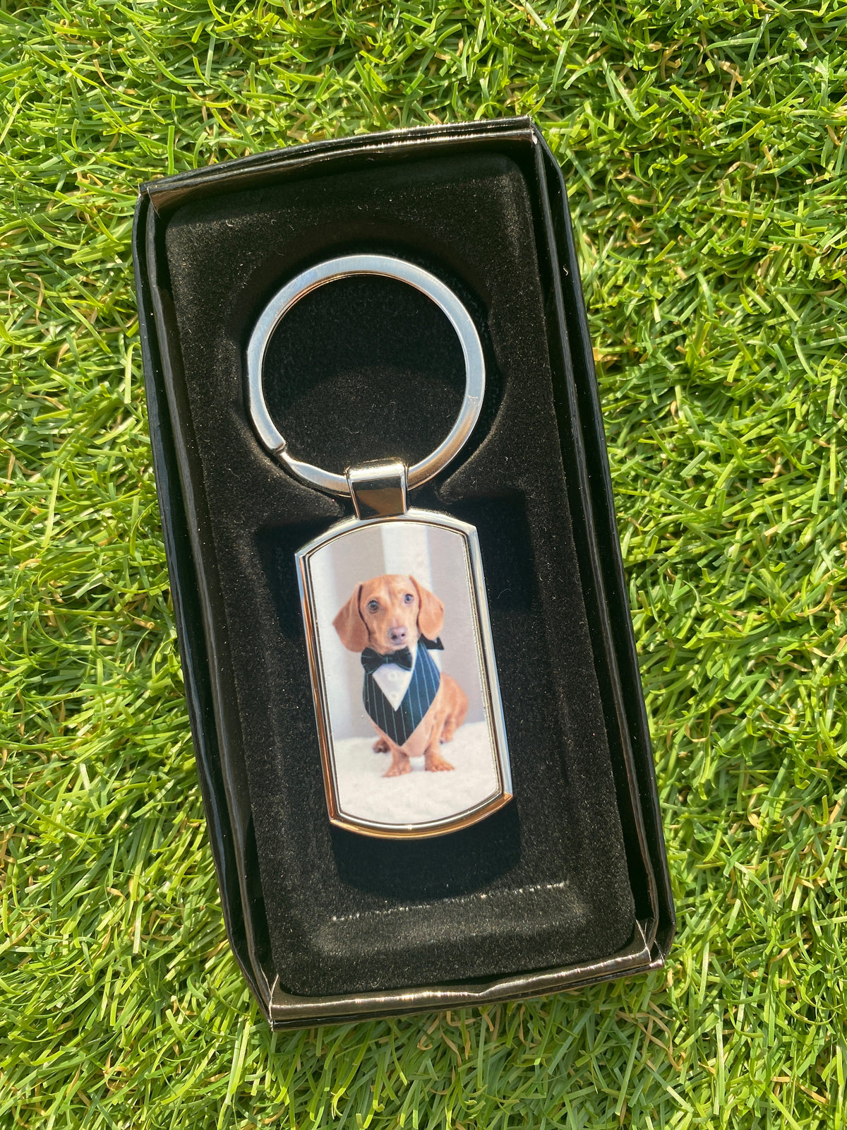 A custom photo keychain with a silver metal split ring and a personalized rectangular tag, displayed inside an open black gift box on a green grass background.
