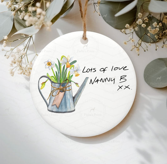 Round ceramic ornament with a watering can and flowers illustration, text 'Lots of love Nanny B xx' on a white background.