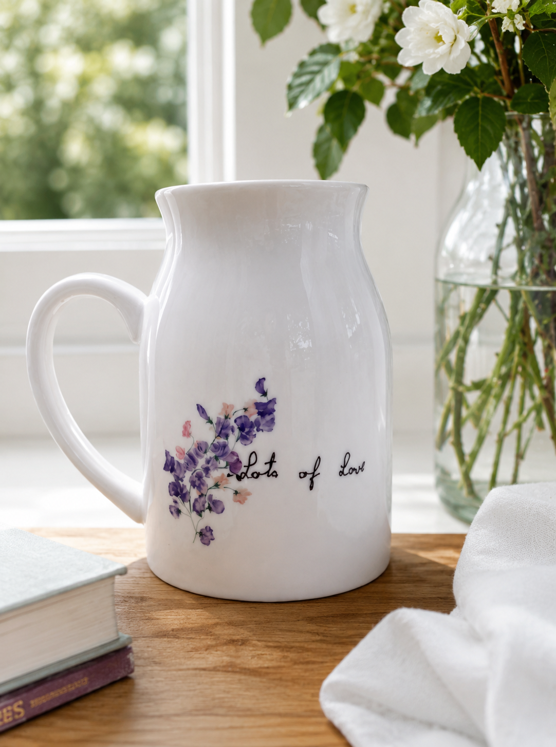 White pitcher with floral design and text on a wooden surface with a window and plant in the background