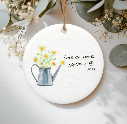 Round ceramic ornament with a watering can and flowers illustration, text  on a white background.