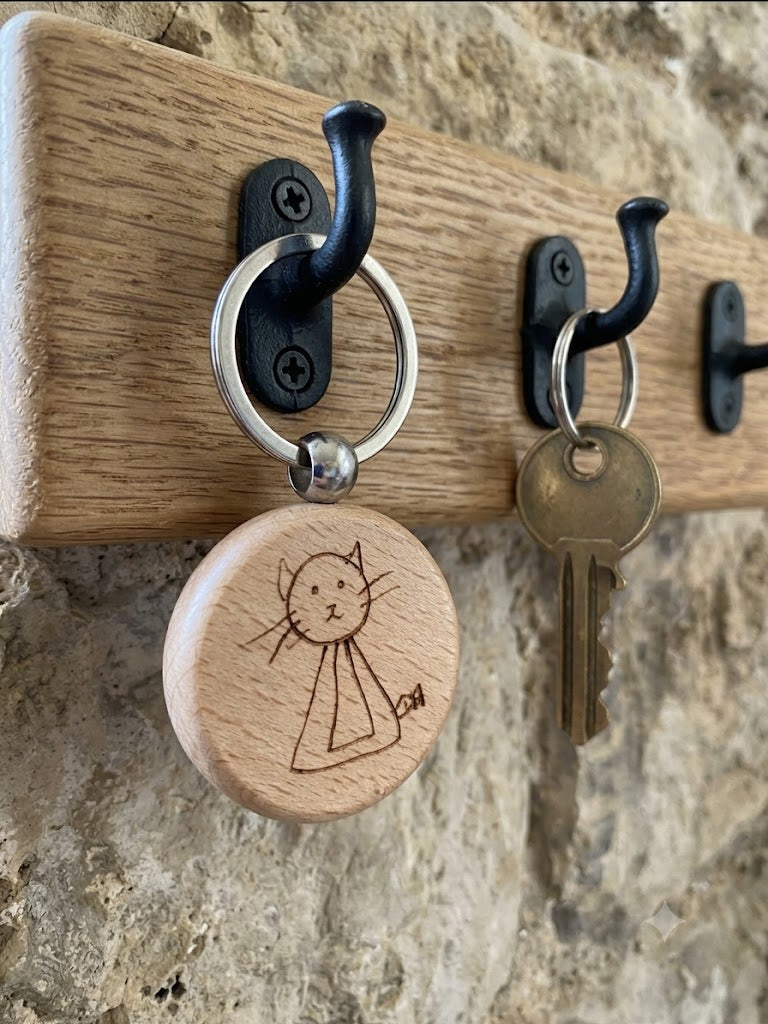 A close-up of a wooden wall-mounted key rack with black metal hooks. One hook holds a round natural wood key tag engraved with a simple cat drawing, and another hook holds a metal key.