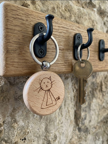A close-up of a wooden wall-mounted key rack with black metal hooks. One hook holds a round natural wood key tag engraved with a simple cat drawing, and another hook holds a metal key.