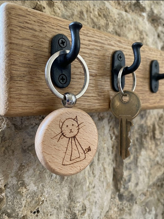 A close-up of a wooden wall-mounted key rack with black metal hooks. One hook holds a round natural wood key tag engraved with a simple cat drawing, and another hook holds a metal key.