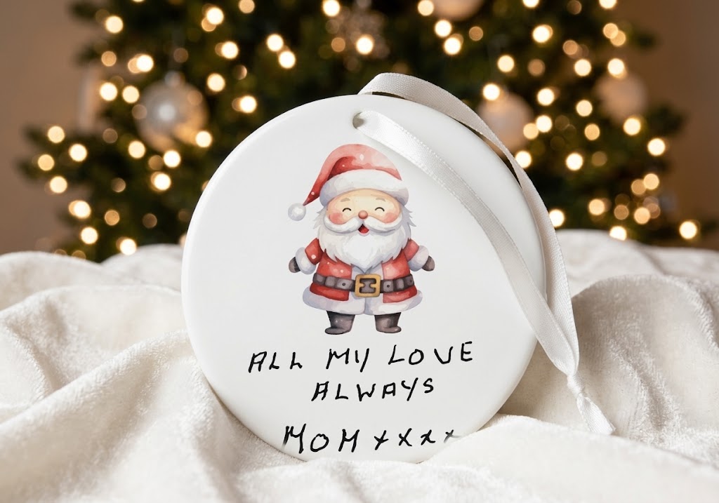 A round white Christmas ornament featuring a cartoon Santa Claus and the text 'ALL MY LOVE ALWAYS MOM XXXX', resting on white fabric with a blurred Christmas tree in the background.