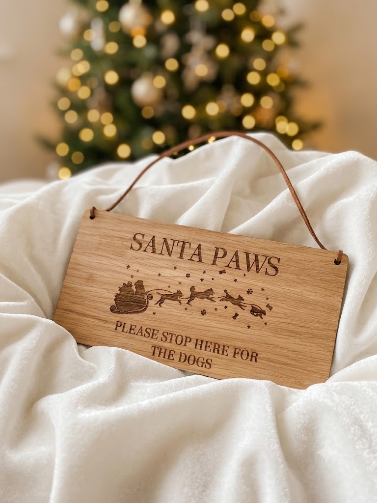 A rectangular wooden hanging sign engraved with 'SANTA PAWS' and 'PLEASE STOP HERE FOR THE DOGS,' plus an illustration of Santa in a sleigh. The sign hangs from a brown cord over white fabric, with a blurred Christmas tree in the background.