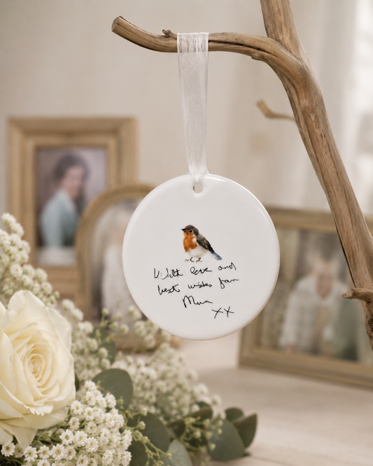 Decorative hanging ornament with a bird illustration and text, placed on a branch with flowers and blurred family photos in the background.