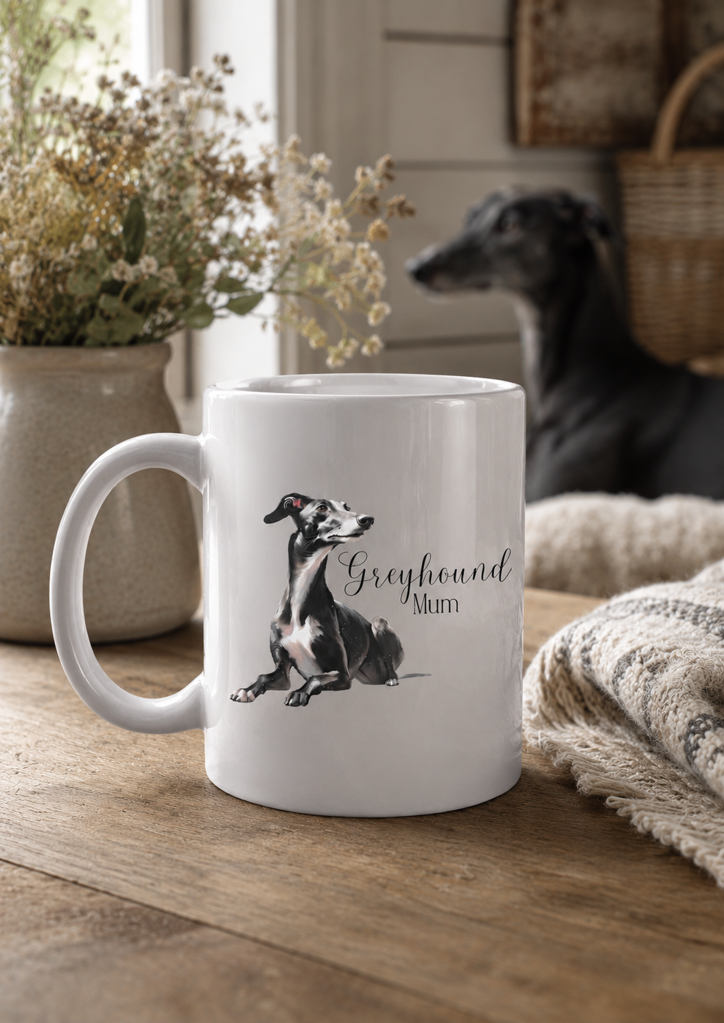 White mug with a dog illustration and 'Greyhound Mum' text on a wooden surface, with a dog and plants in the background.