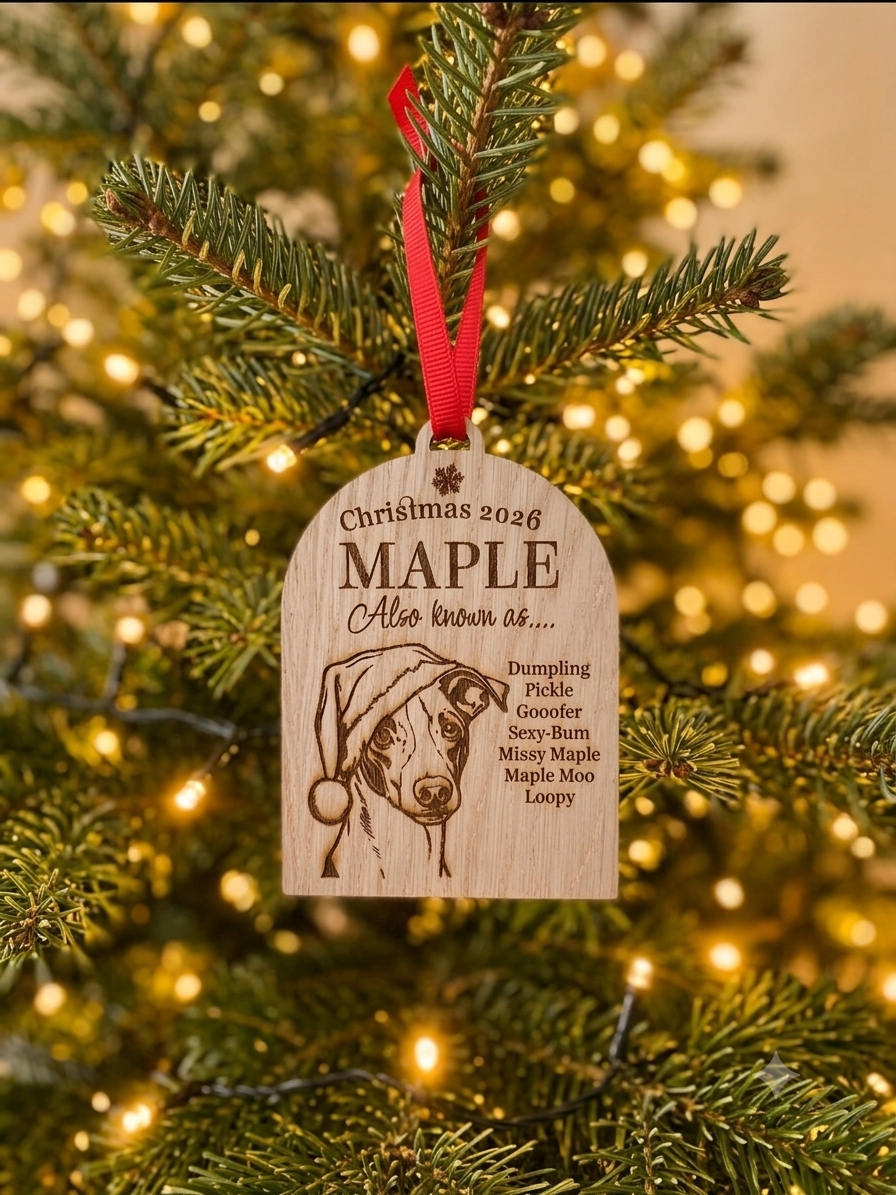 A personalized wooden Christmas ornament hanging from a pine tree branch with warm string lights in the background. The ornament is engraved with a dog portrait and the text 'Christmas 2026 MAPLE Also known as.