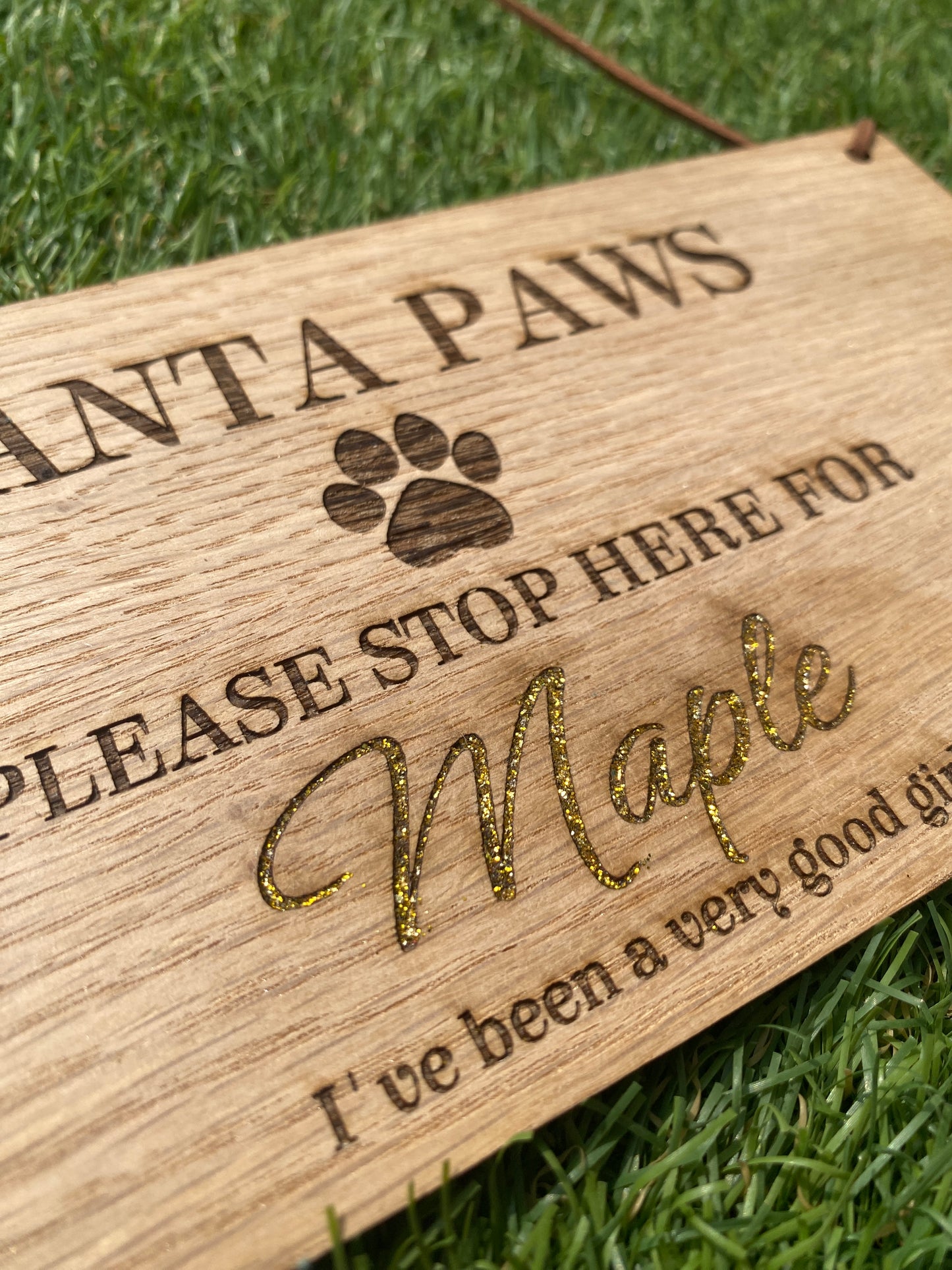 A close-up of a wooden engraved plaque with a paw print and the text 'Please Stop Here For [glittery script name]', placed on green grass.