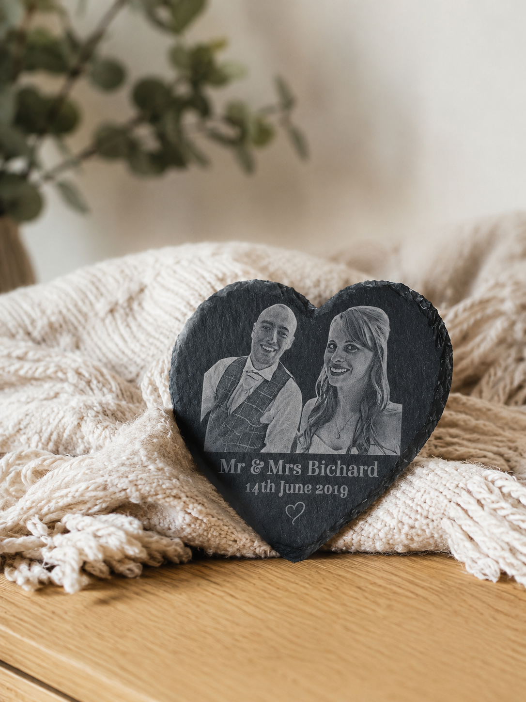 Heart-shaped engraved stone with a photo of a couple and text, placed on a textured surface with a blurred plant in the background.