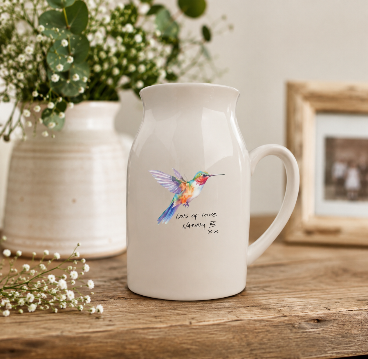 White mug with a colorful hummingbird design on a wooden surface with flowers and a photo frame in the background.