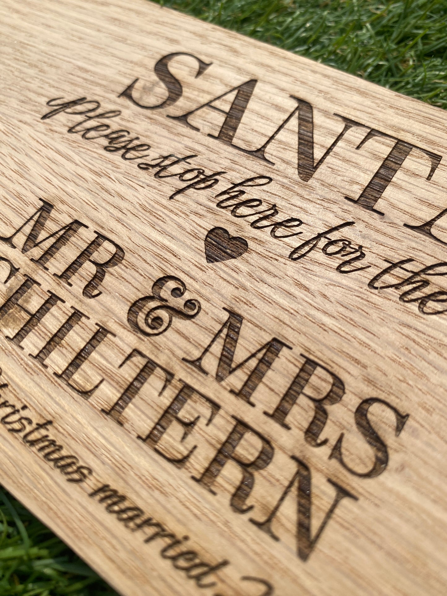 A close-up of a personalized wooden plaque engraved with 'SANTA', 'MR & MRS WHITERN', and 'Please stop here for the Christmas married' text, resting on green grass.