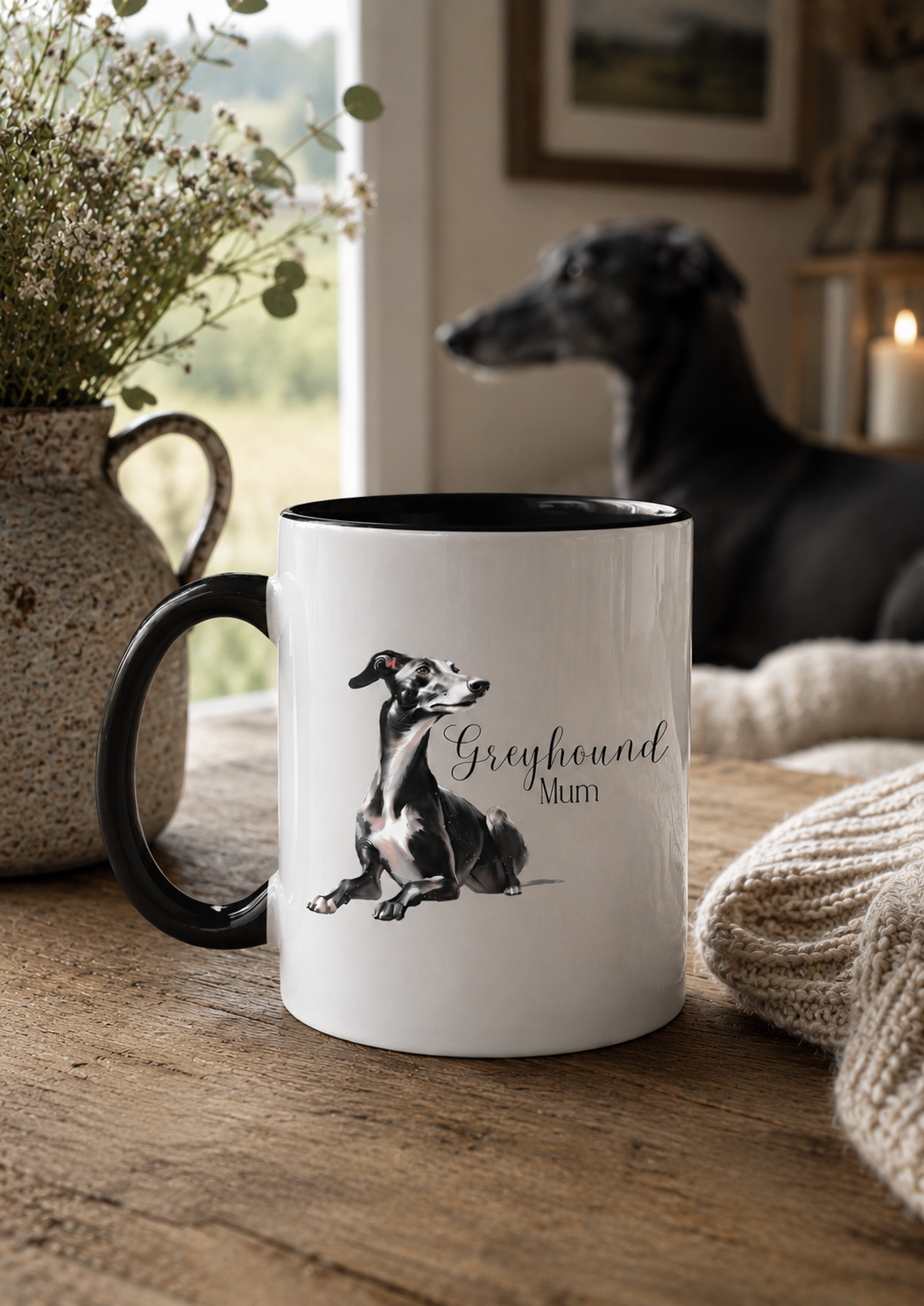 White mug with black interior featuring a greyhound illustration and text, placed on a wooden surface with a dog in the background.