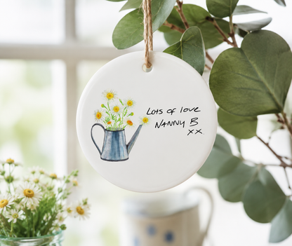Round ceramic ornament with watering can illustration and text, hanging on a plant.