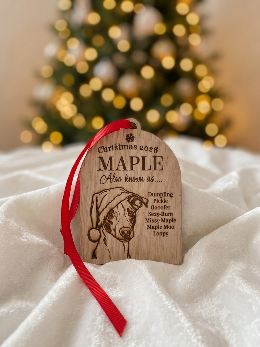 A personalized wooden Christmas ornament featuring an engraved dog portrait and red ribbon, displayed on white fabric with a blurred Christmas tree in the background.