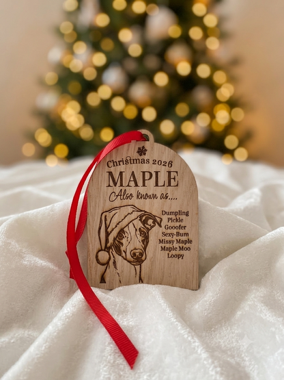 A personalized wooden Christmas ornament featuring an engraved dog portrait and red ribbon, displayed on white fabric with a blurred Christmas tree in the background.