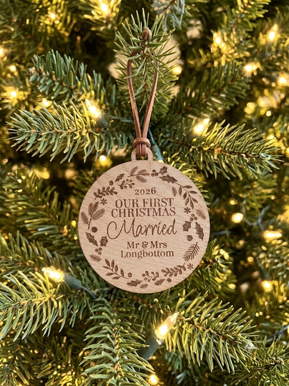 A personalized round wooden Christmas ornament hanging on a lit Christmas tree. The natural wood disc features an engraved brown floral wreath border and the text '2026 OUR FIRST CHRISTMAS Married Mr & Mrs Longbottom'.