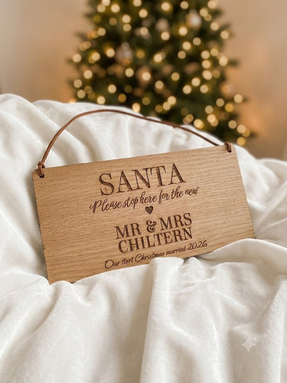 A personalized wooden hanging sign engraved with 'SANTA Please stop here for the new MR & MRS CHILTERN Our first Christmas married 2026', resting on white draped fabric with a blurred Christmas tree in the background.