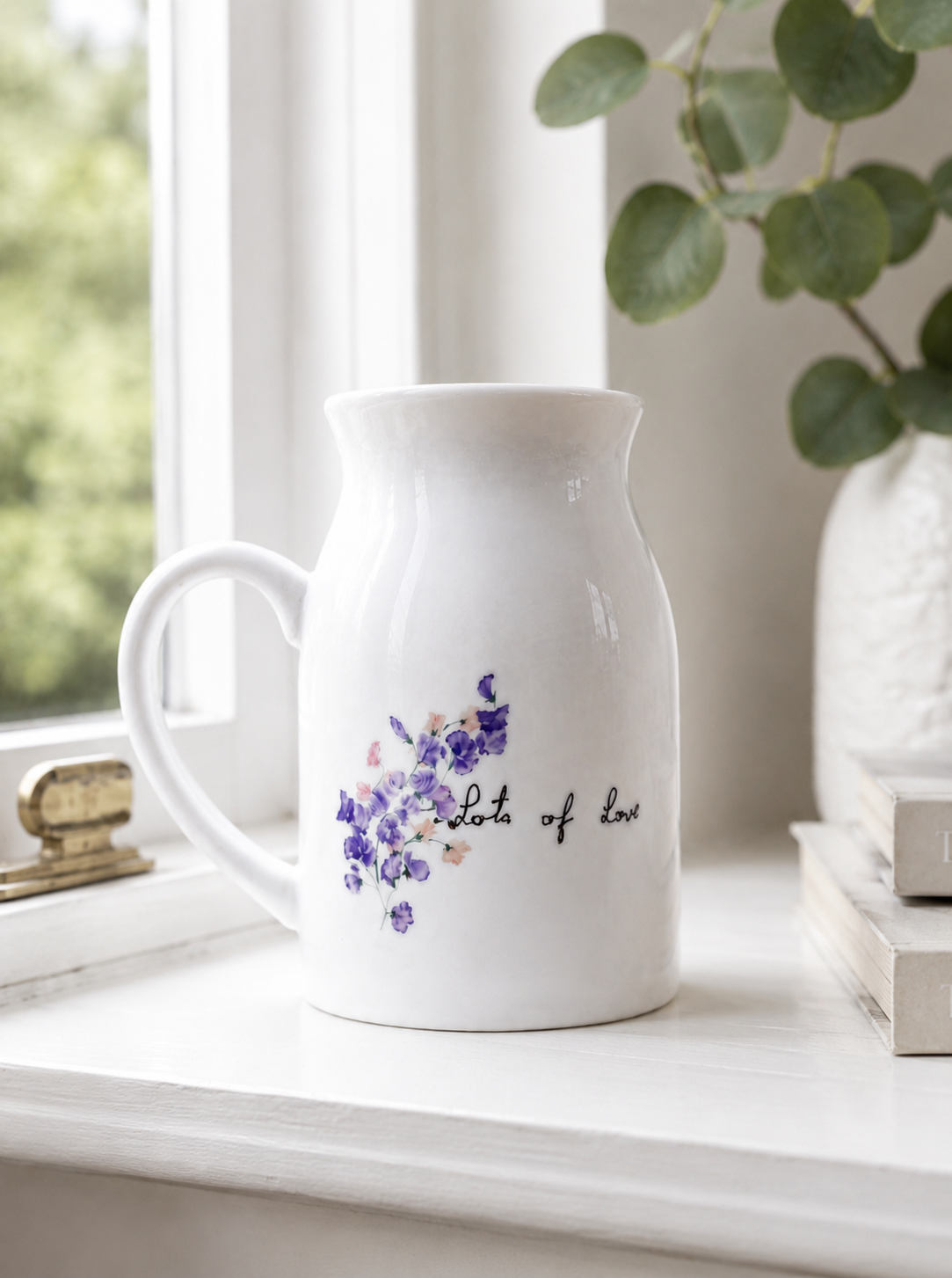 White ceramic pitcher with floral design and text on a windowsill with a plant in the background