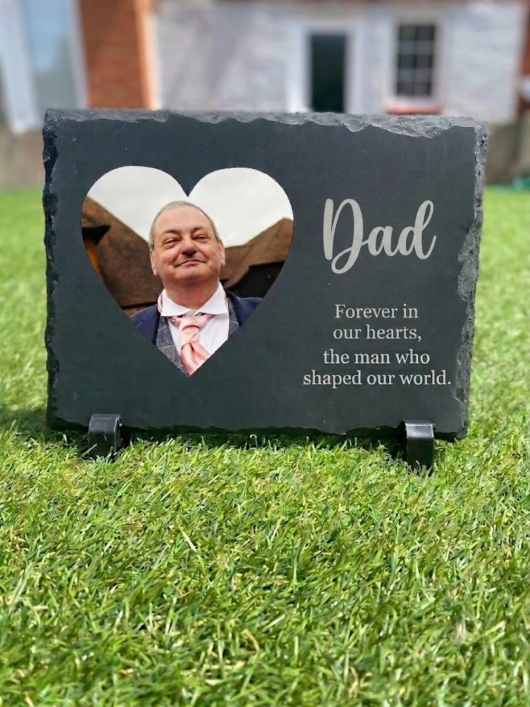 A personalized dark slate memorial plaque on a small stand, placed on green grass outdoors. The plaque has a heart-shaped cutout showing a portrait of a man, with the word 'Dad' and the engraved remembrance quote 'Forever in our hearts, the man who shaped our world.