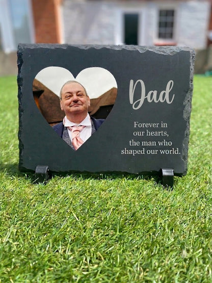 A personalized dark slate memorial plaque on a small stand, placed on green grass outdoors. The plaque has a heart-shaped cutout showing a portrait of a man, with the word 'Dad' and the engraved remembrance quote 'Forever in our hearts, the man who shaped our world.