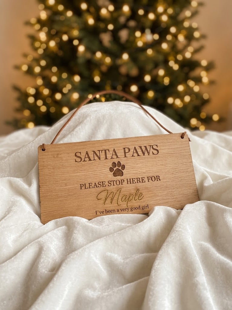 A personalized wooden hanging sign engraved with 'SANTA PAWS PLEASE STOP HERE FOR Maple I've been a very good girl' and a paw print, displayed on white draped fabric with a blurred Christmas tree in the background.