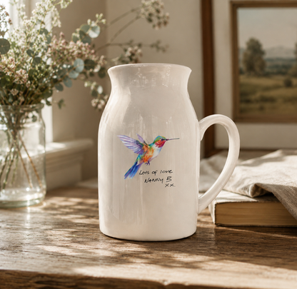 White ceramic pitcher with a colorful hummingbird design on a wooden surface with flowers and a book in the background.