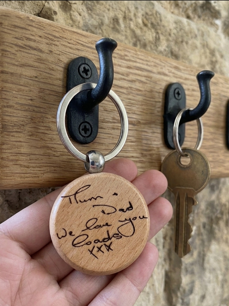 A hand holds a round engraved wooden keychain in front of a wall-mounted wooden coat rack with black hooks. The keychain has a silver split ring and a light natural wood tag with black handwritten 