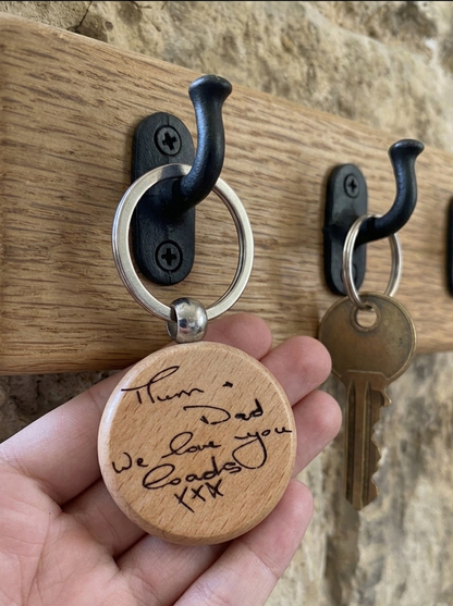 A hand holds a round engraved wooden keychain in front of a wall-mounted wooden coat rack with black hooks. The keychain has a silver split ring and a light natural wood tag with black handwritten 