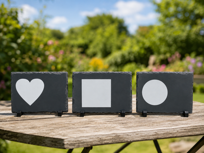 Three rectangular black slate signs on a wooden table in a garden, featuring white cutout shapes of a heart, a square, and a circle, with a blurred green garden background.