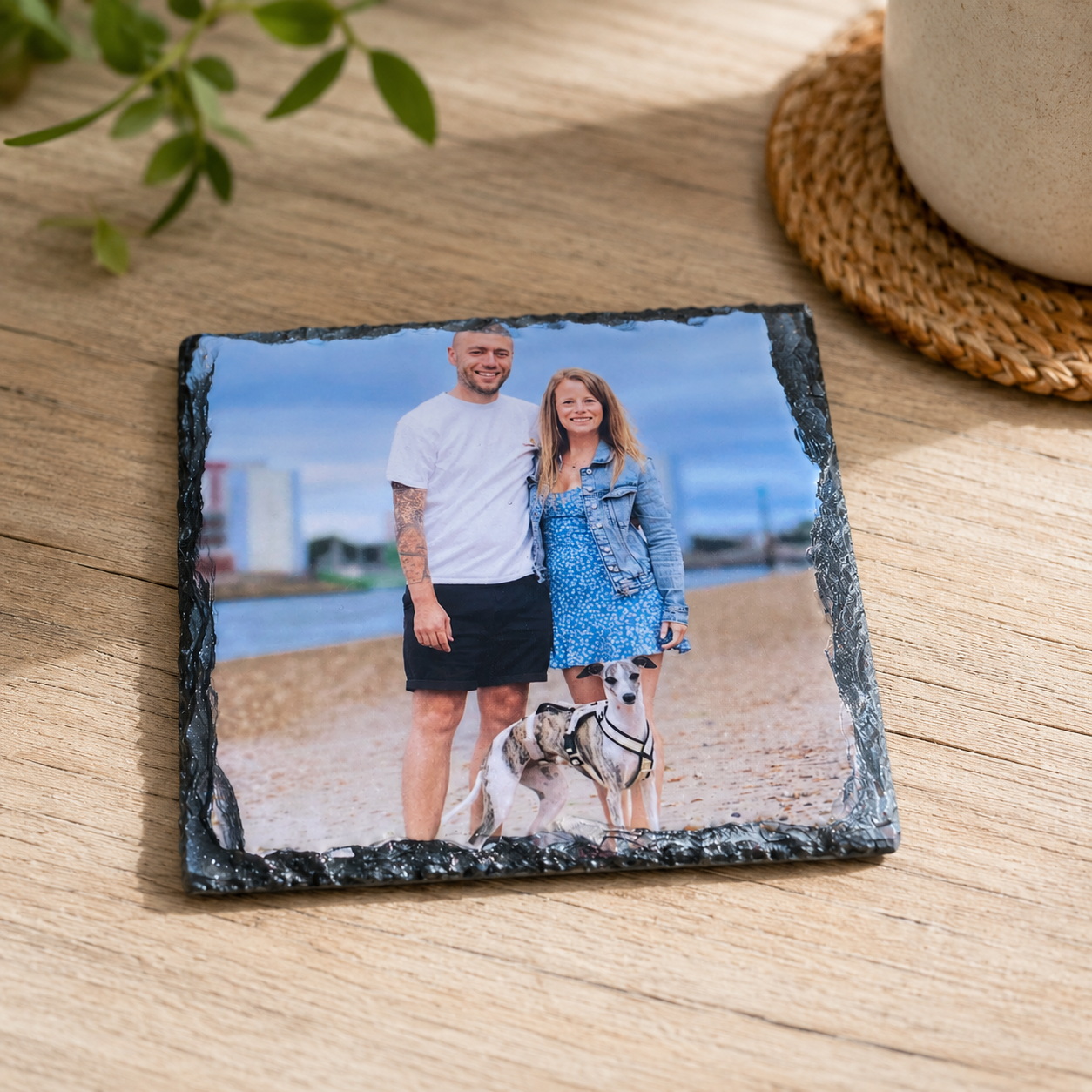 A square slate photo coaster with a black natural stone border, displaying a printed photograph of a couple and their dog on a beach, placed on a light wood tabletop.