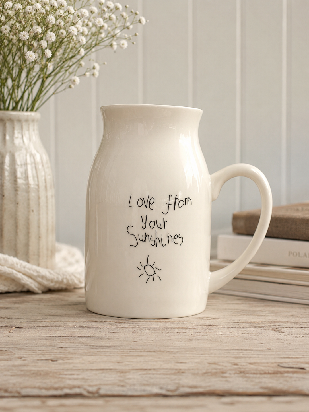 White ceramic mug with 'Love from your Sunshines' text on a wooden surface with a vase of flowers and books in the background.