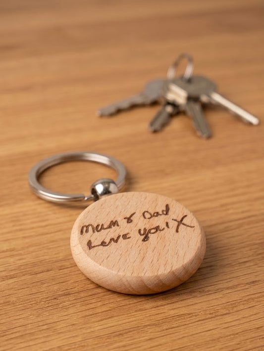 A personalized round wooden keychain engraved with 'Mum & Dad Love you X' in brown handwriting, attached to a silver metal split ring. The keychain is displayed on a warm wood-grain tabletop next to a set of keys in the background.