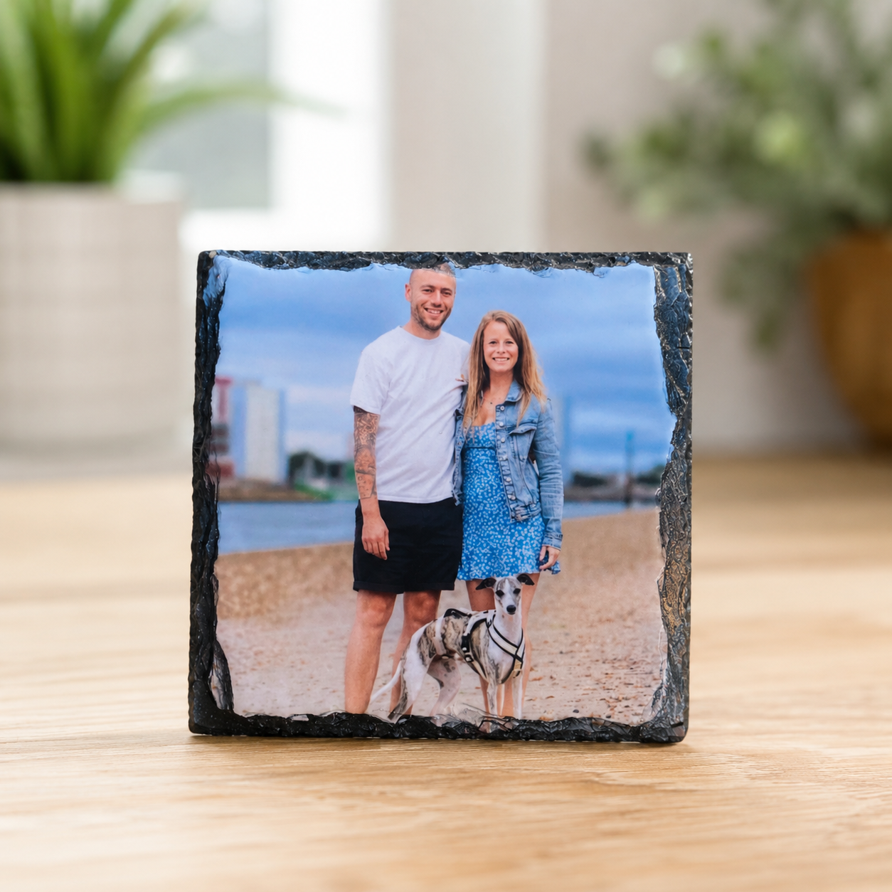 A square black slate photo plaque standing on a wooden table, displaying a printed photograph of a couple and their dog.