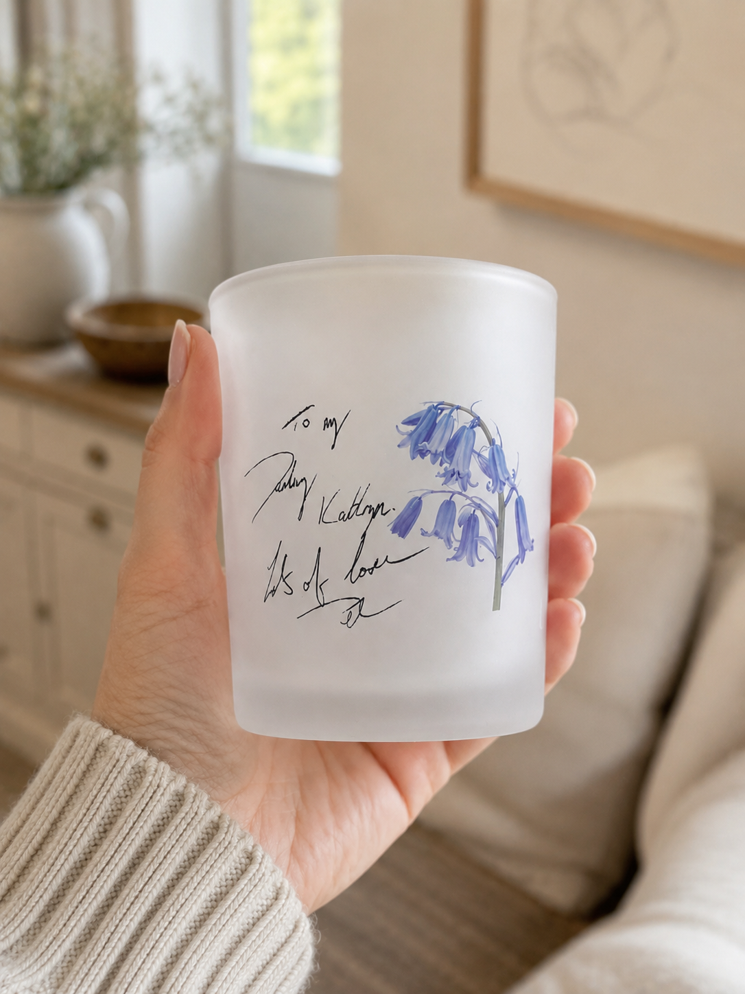 Frosted glass candle holder with blue floral design held in a hand in a home setting.