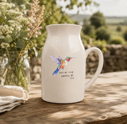 White mug with a colorful hummingbird design on a wooden table outdoors.