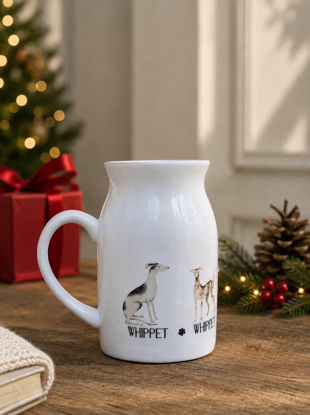 A white ceramic milk jug mug featuring illustrations of a whippet dog and a deer with the word 'WHIPPET' printed below, set on a wooden table with a blurred Christmas background.