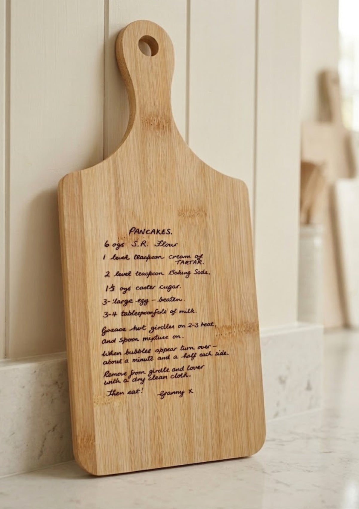 A rectangular wooden serving board with a rounded handle and a printed recipe for pancakes, leaning against a white paneled wall on a light marble surface.