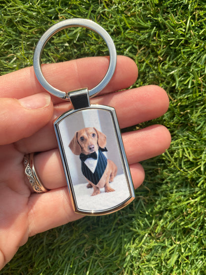 A silver metal keychain with a custom printed photo of a dachshund wearing a tuxedo, held in a person's hand against a background of green grass.
