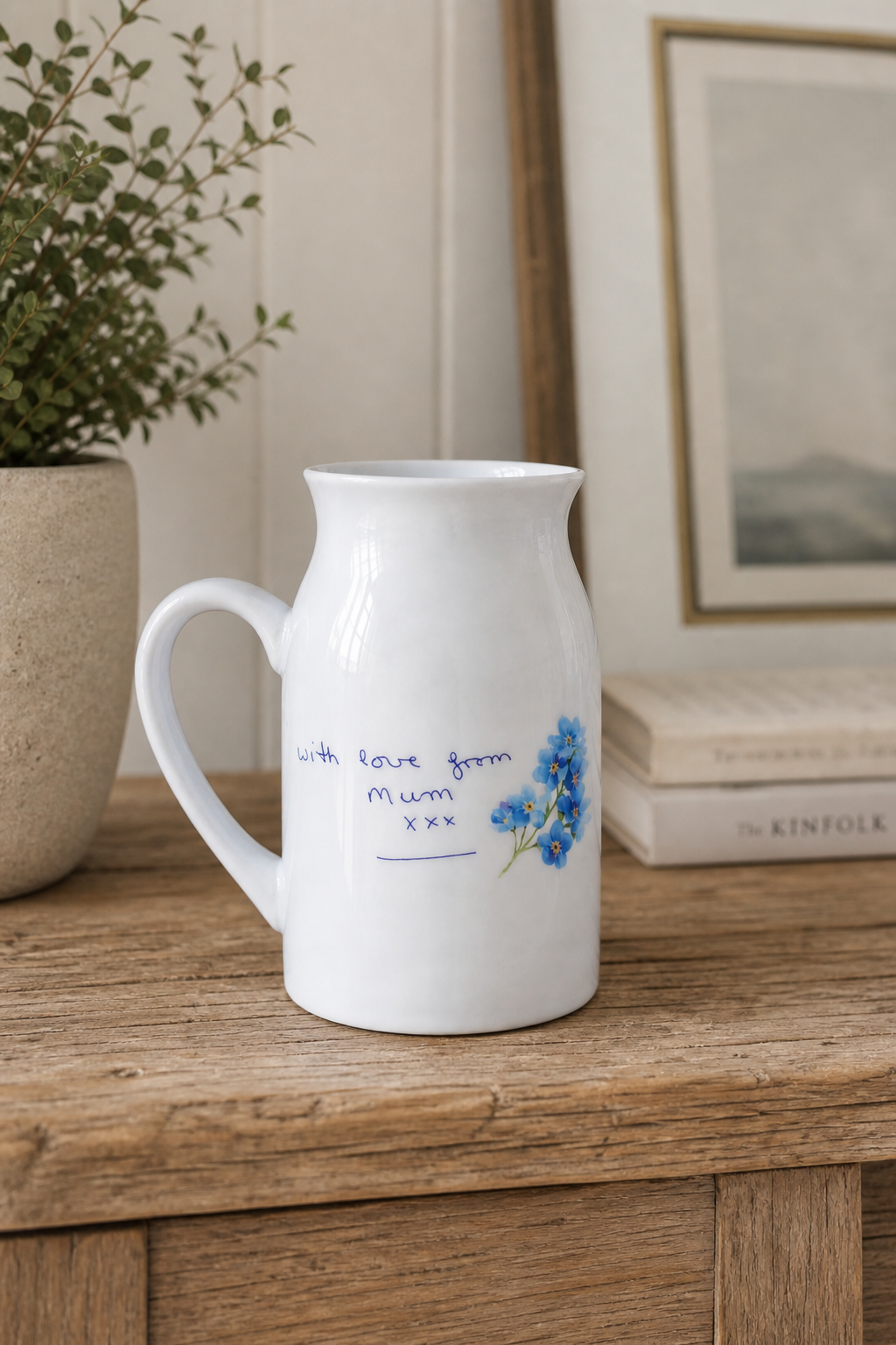 White ceramic pitcher with floral design and text on a wooden surface, with a plant and books in the background.
